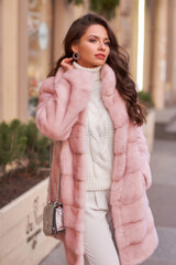 Young elegant pretty woman with long wavy hear wearing white trousers and pullover and pink fur...