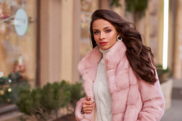 Young elegant pretty woman with long wavy hear wearing white trousers and pullover and pink fur coat walking city street at winter. Stylish female model