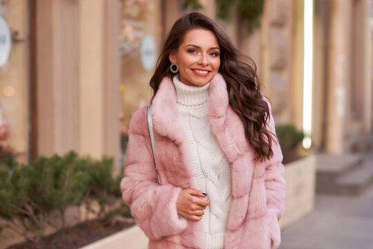 Happy smiling woman outdoor winter portrait. Pretty young rich female in white casual costume and pink fur coat with long wavy hair and makeup walking city street - Powered by Adobe