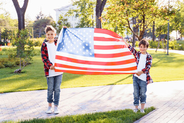 America's USA Independence Day. Two cute brothers guy americans standing in a city park on skateboards and holding flag of USA. Stretching fabric and shouting exiting with joy in flannel shirts