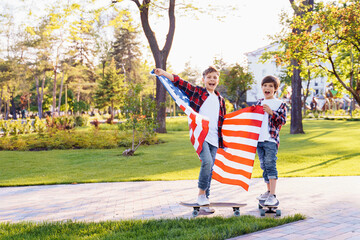 America's USA Independence Day. Two cute brothers guy americans standing in a city park on skateboards and holding flag of USA. Stretching fabric and shouting exiting with joy in flannel shirts