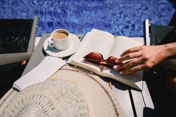 Coffee and book at the pool side.Female Sits Near By Pool at Summer Time Reading Book Drink...