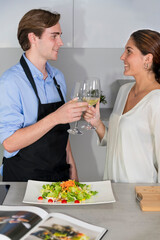 Smiling young couple having a toast with glasses of white wine next to a kitchen counter with a salad and an out of focus recipe book