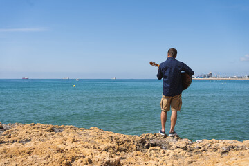 Young man with a printed shirt on his back playing a Spanish guitar standing in front of the sea