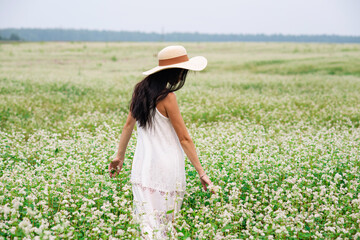 A girl with dark hair in a white dress and a straw hat walks on a white flower field. Summer rural landscape. Tender brunette portrait