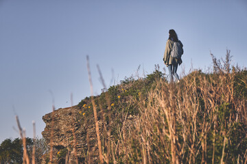 back view of a girl walking along a trail through the grass at the edge of a cliff with sky background. Tourism and outdoor concept.