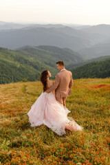 The bride and groom are standing with their backs and hugging. Sunset. Wedding photo on a background of autumn mountains. A strong wind inflates hair and dress.