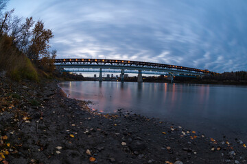THE HIGH LEVEL BRIDGE AT 100, in a beautiful sunrise with shades of blue, yellow and red lights, over a river of blue water with reflections and swept clouds, stones and yellow leaves in edmonton city