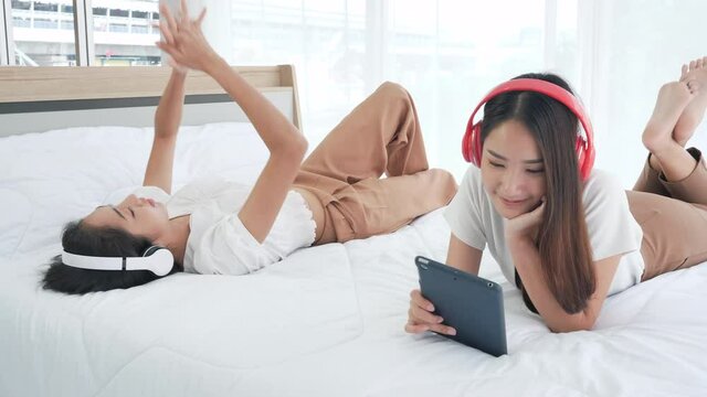 Two Asian teenage girls happy in headphones using Digital tablet lying listening to music on bed at home. woman relax, lifestyle, youth and pleasure concept. Dolly slider shot