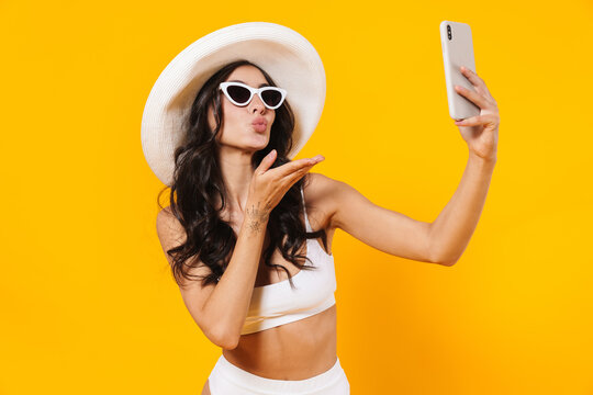 Image of elegant cute woman in swimsuit and hat blowing air kiss and using cellphone