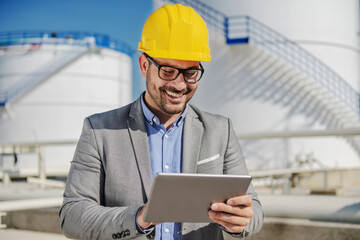 Portrait of smiling satisfied caucasian unshaven businessman with helmet on head using tablet to check prices on stock for oil. Refinery exterior. In background are tanks with oil.