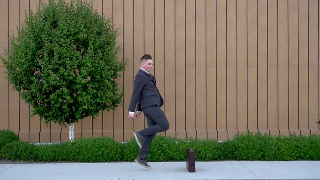 A young businessman in a suit is dancing. Modern youth flex. A man dances with joy against the background of a building with a flowerbed. Slow motion