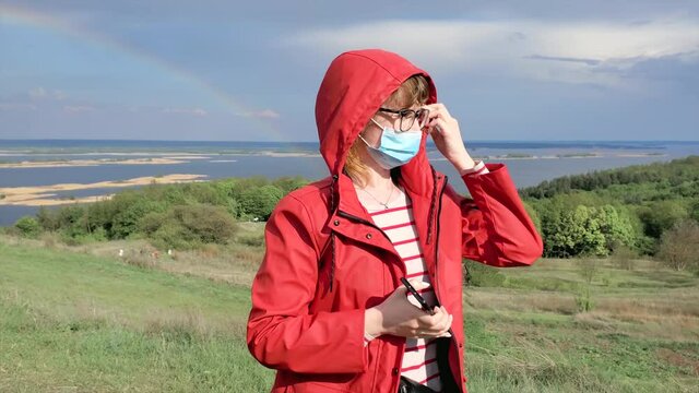Woman in a red jacket and a protective medical mask standing against a rainbow and a stormy sky