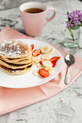 A stack of pancakes with a pink tea mug and  fresh strawberries on a white plate on a light background. A small bouquet of lilacs in a glass vase stands next to the Breakfast.