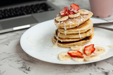 A stack of pancakes with fresh strawberries on a white plate on a light background. There is a laptop next to it. Work at home. Horizontal orientation. Homemade american pancakes. Copy space.