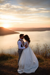 The bride and groom are hugging on the background of the lake during sunset.