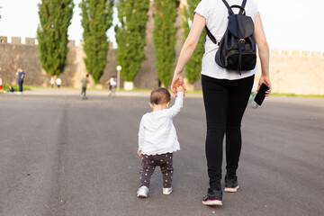 Fototapeta premium First steps of little girl. Mother and her toddler holding hands and walking away together on playground.