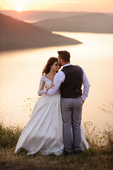 The bride and groom are hugging on the background of the lake during sunset.