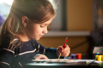 Side view of charming little caucasian smiling schoolgirl writes while sitting at table. Little cute girl draws picture. Concept of teaching children during self-isolation