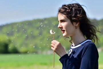 Portrait of a young girl blowing dandelion flowers in the background with trees
