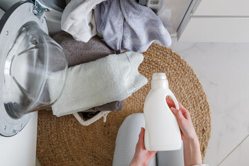 Woman holds bottle with liquid laundry detergent before washing clothes. Washer and clothes ready...