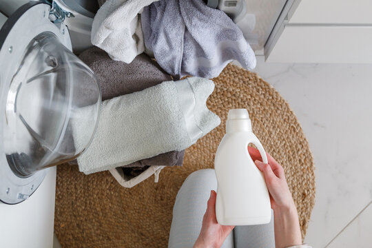 Woman Holds Bottle With Liquid Laundry Detergent Before Washing Clothes. Washer And Clothes Ready For Laundry. View From Above