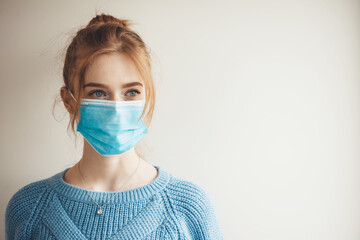 Ginger girl with freckles wearing medical mask and knitted blue sweater is posing on a white studio wall with free space