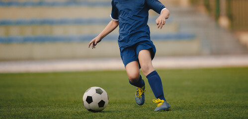 Junior level football player running  with a ball. Footballer in soccer cleats and jersey blue uniform kicking classic soccer ball on grass pitch. Sports arena in the background