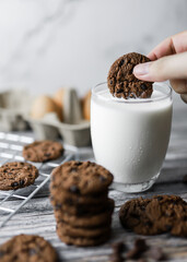 a hand dipping cookie in a tall glass of milk with a stack of homemade chocolate chip cookies