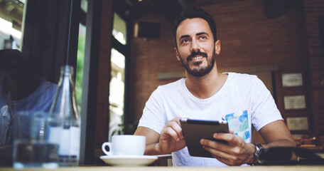 Happy man proprietor of cozy cafe rejoice in own success while sitting with touch pad at dining table, 