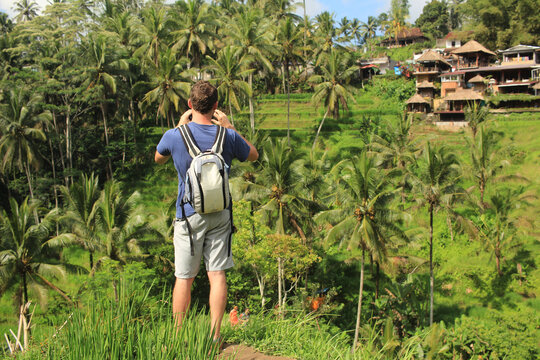 Caucasian tourist with backpack standing in lush green rice fields and palm trees taking photos on a bright sunny day in Bali Indonesia