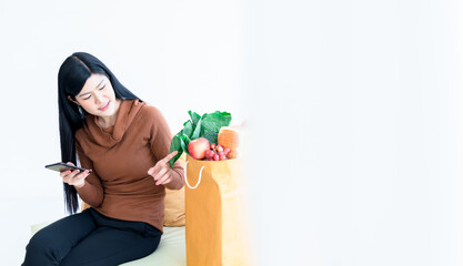 Attractive Asian woman Holding a cell phone And checking the product Which is fresh vegetables and fruits which she ordered online, concept to new normal and shopping online