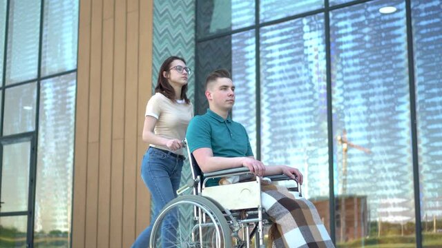 A young woman is carrying a young man in a wheelchair. An assistant carries a disabled person in a wheelchair along the street. Special transport for the disabled.