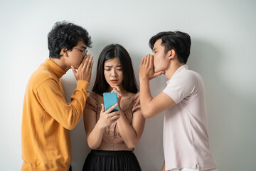 two young men whispering to a young girl using a smart phone while standing against an isolated background