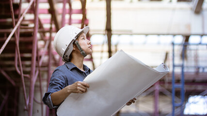 Female engineer wearing safety helmet while holding the blueprint on her hands. Woman engineering inspect the site with blue and pink scaffolding background.