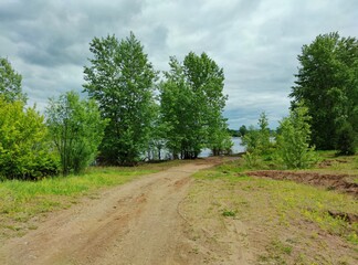Fototapeta na wymiar country fishing road leading to green trees by the river against a cloudy sky before the rain