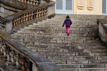 Unrecognisable little girl, modern-day lonely princess walking up majestic curved stairs of empty...