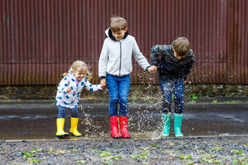 Three children, toddler girl and two kids boys wearing red, yellow and green rain boots and walking...