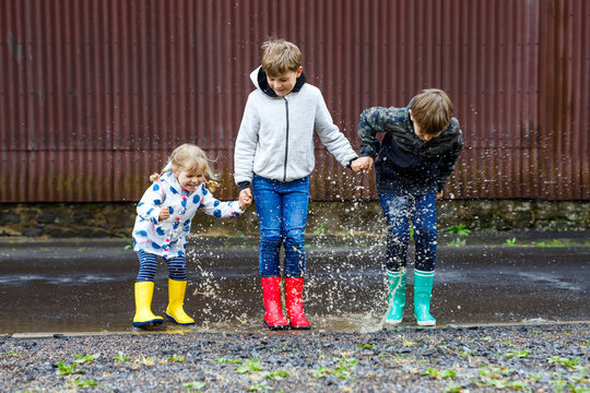 Three children, toddler girl and two kids boys wearing red, yellow and green rain boots and walking during sleet. Happy siblings jumping into puddle. Having fun outdoors, active family