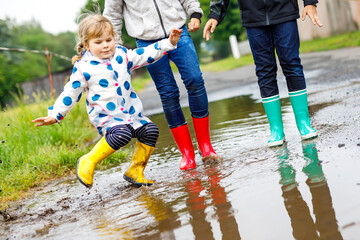 Three children, toddler girl and two kids boys wearing red, yellow and green rain boots and walking during sleet. Happy siblings jumping into puddle. Having fun outdoors, active family