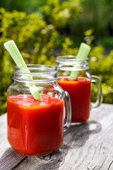Two glasses of tomato juice with parsley and celery decorations, stand in the open air on wooden boards, the rays of the sun shine, shallow depth of field, selective focus. The concept of natural