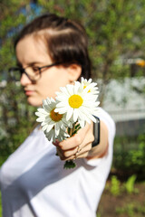 A young girl with glasses holds out a small bouquet of daisies.