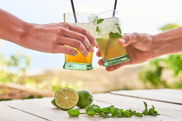 Two hands with mojitos toasting, with the beach in the background. Concept of drink for summer. - 356191913