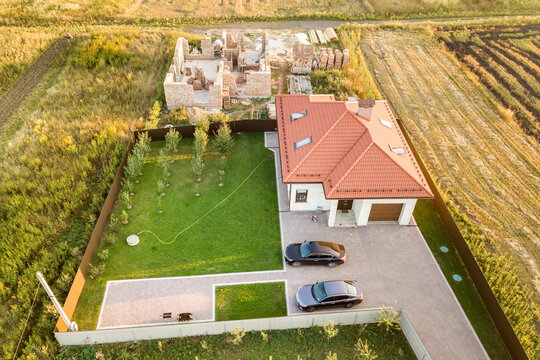 Top down aerial view of two private houses, one under construction with concrete foundament and brick walls and another finished with red tiled roof.