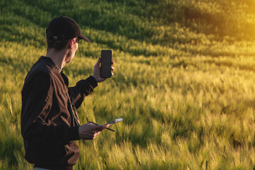 Young agronomist in cap takes notes in a notebook on a green agricultural field during sunset