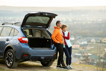 Happy couple standing together near a car with open trunk enjoying view of rural landscape nature. Man and woman leaning on family vehicle luggage compartment. Weekend travel and holidays concept. © bilanol