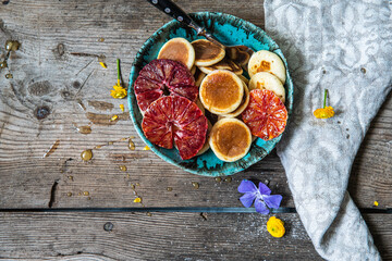 Ceramic bowl with mini pancakes, blood orange slices and honey on old wooden table.