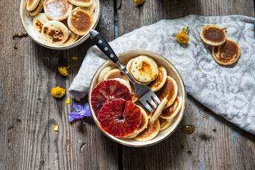 Ceramic bowl with mini pancakes, blood orange slices and honey on old wooden table.