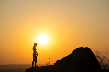 Silhouette of a woman hiker standing alone near big stone at sunset in mountains. Female tourist on high rock in evening nature. Tourism, traveling and healthy lifestyle concept.