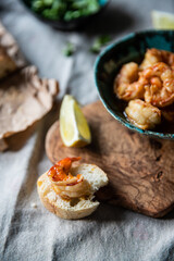 Slice of baguette with roasted red shrimp on wooden board with turquoise bowl with garlic shrimps and lemon quarters on linen tablecloths.